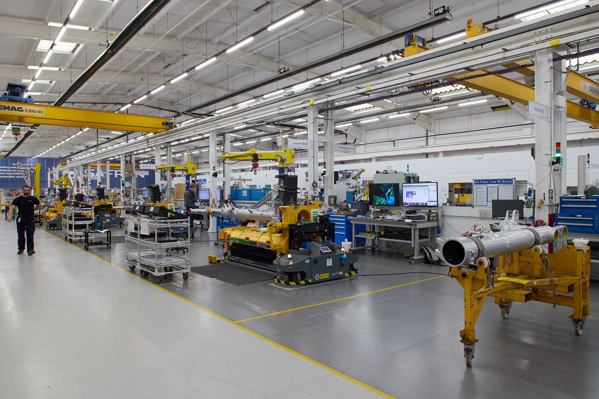 Safran Landing Systems inside of large factory with clear floors and high ceiling and strip lighting there are yellow steel  metal machines and blue machines