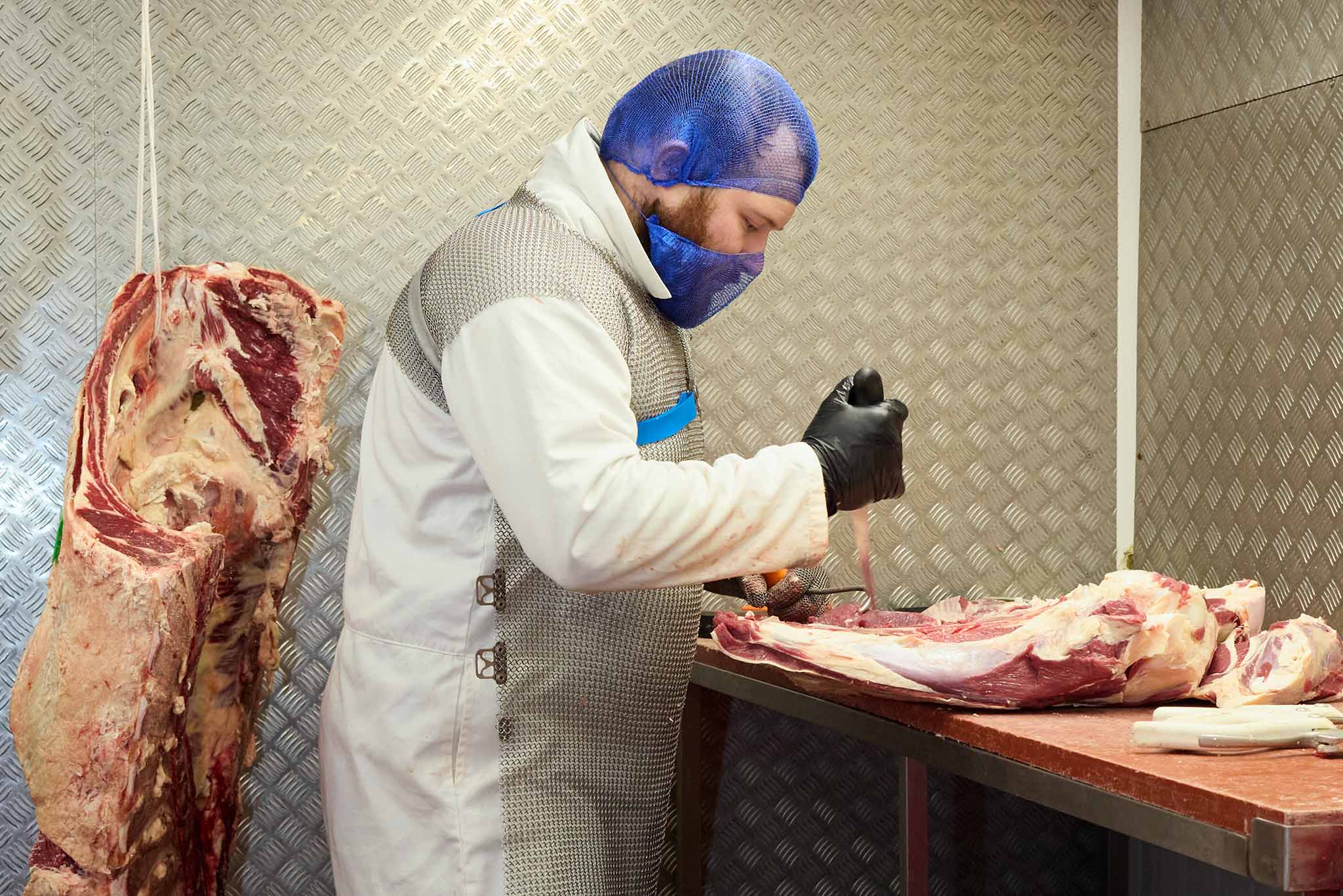 Swaledale Butchers man butchering cut of meat with large animal carcass hanging behind him
