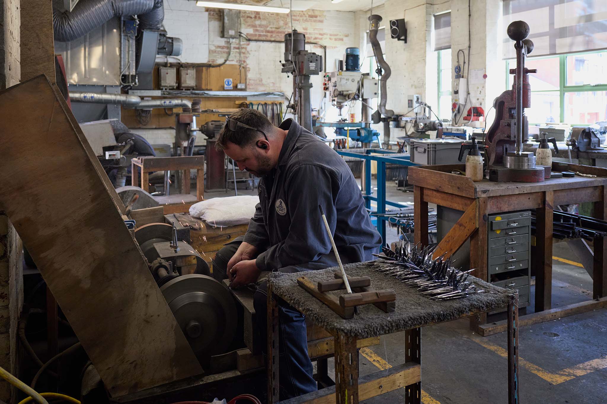 Ernest Wright in workshop man in overalls sitting working at grinding wheel sparks flying to his left there is a table with multiple parts of schissors