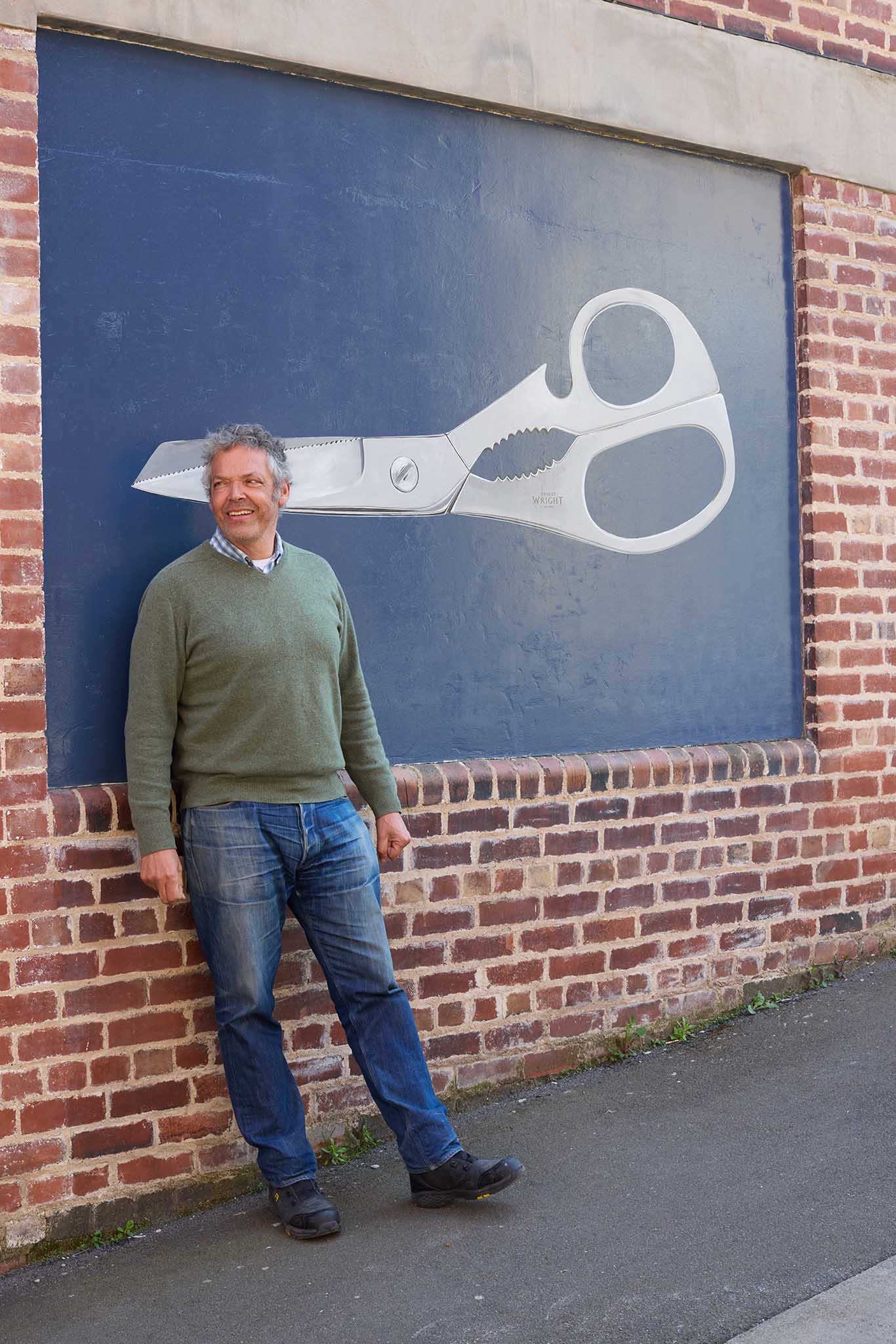 Ernest Wright man in jeans and green jumpers leaning against a brick wall with large bright blue picture sign on wall with large pair of grey scissors painted