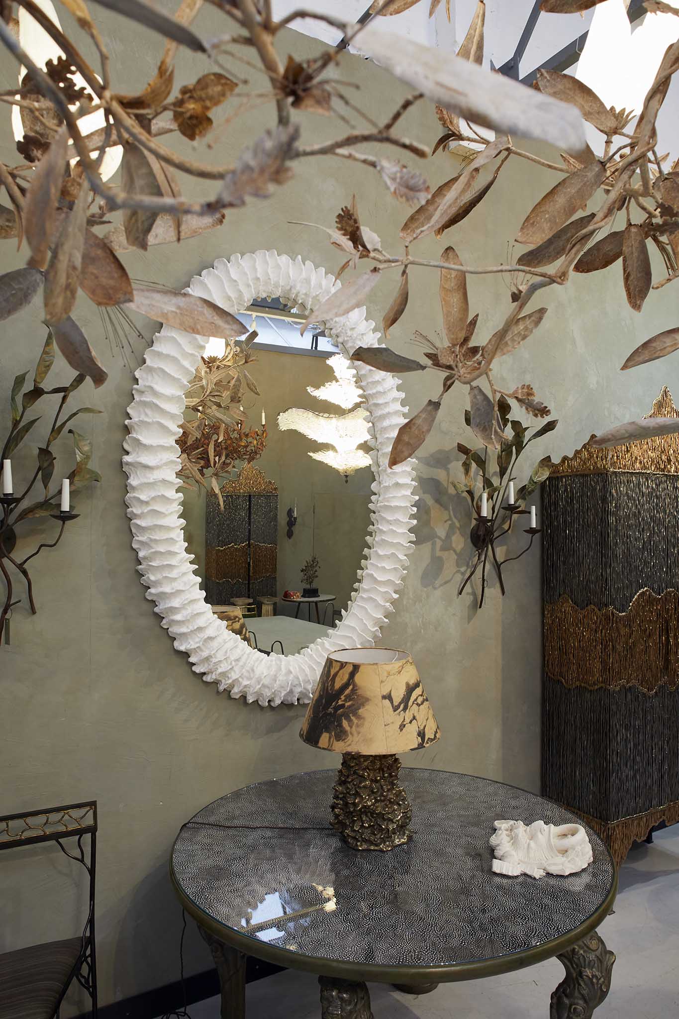 Cox London inside showroom circular table with lamp on top there is a white circular mirror on the wall and above is a delicate metal leaf golden sculplture