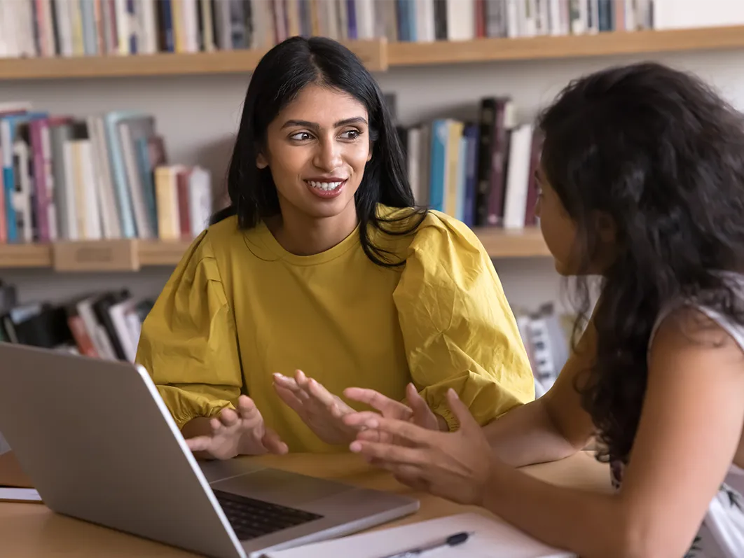 Young female apprentice in an office setting and chatting to employer engaged in conversation whilst on laptops.