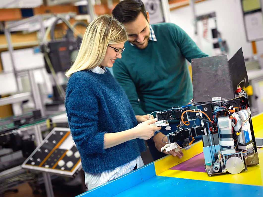 Young female HTQ student working alongside employer in an electronics lab, wiring some machinery.