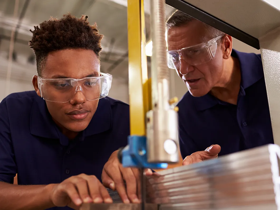 Young male wearing protective goggles during T Level, working alongside an employer and operating a large saw