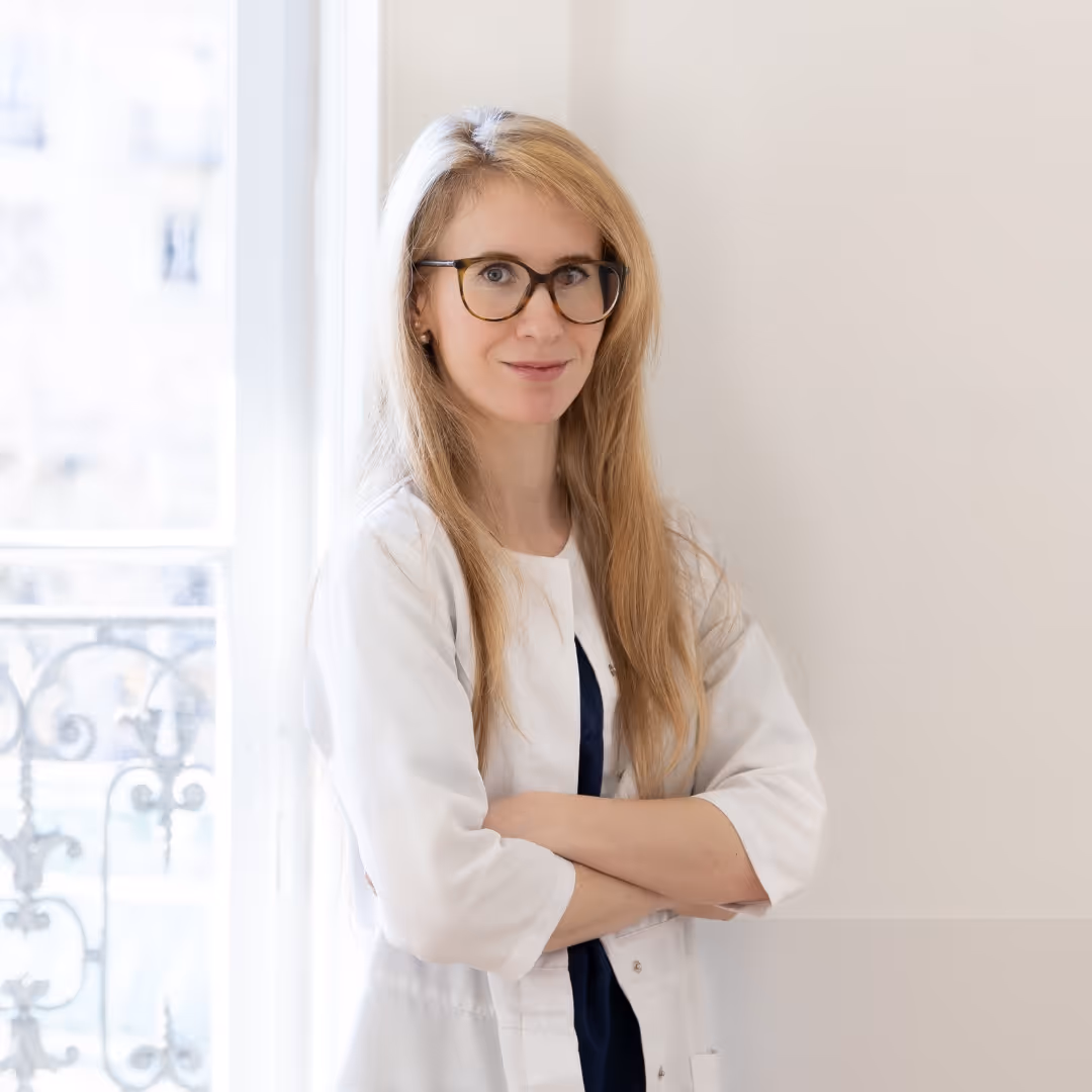 Portrait du Dr Caroline De Lorenzi, dermatologue, aux cheveux blonds longs, lunettes écaille, blouse blanche, bras croisés, devant une fenêtre lumineuse