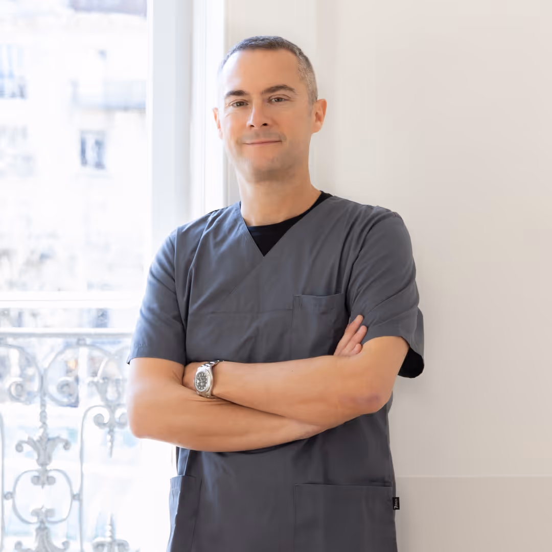 Portrait du Dr Rocco Della Torre, dermatologue, aux cheveux gris courts, tenue soignante grise, bras croisés, stylo en poche, devant une fenêtre avec balcon en fer forgé