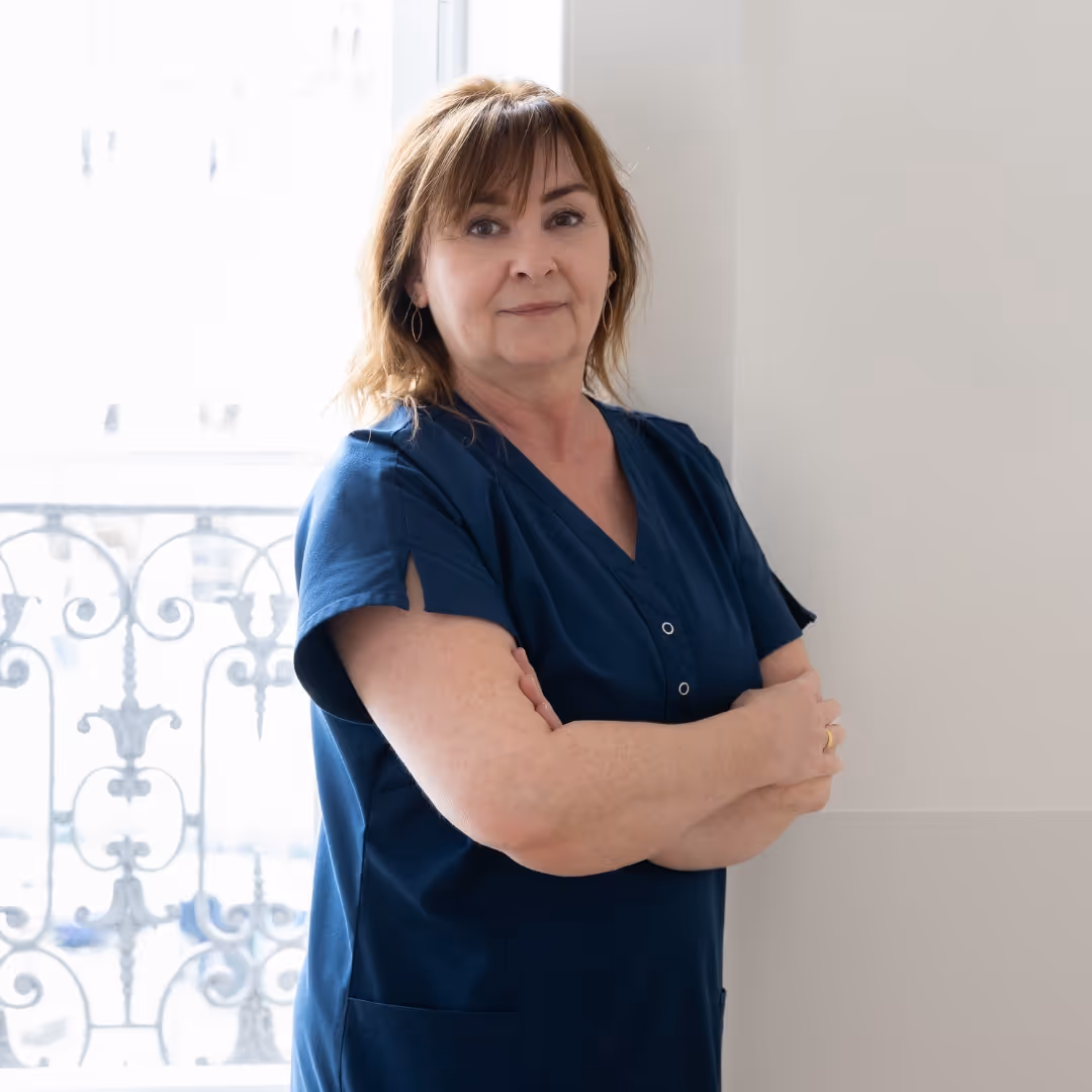 Portrait d'Isabelle Bonvallat Gertsch, esthéticienne, aux cheveux châtains mi-longs avec frange, bras croisés, tenue soignante bleue marine, devant une fenêtre avec balcon en fer forgé