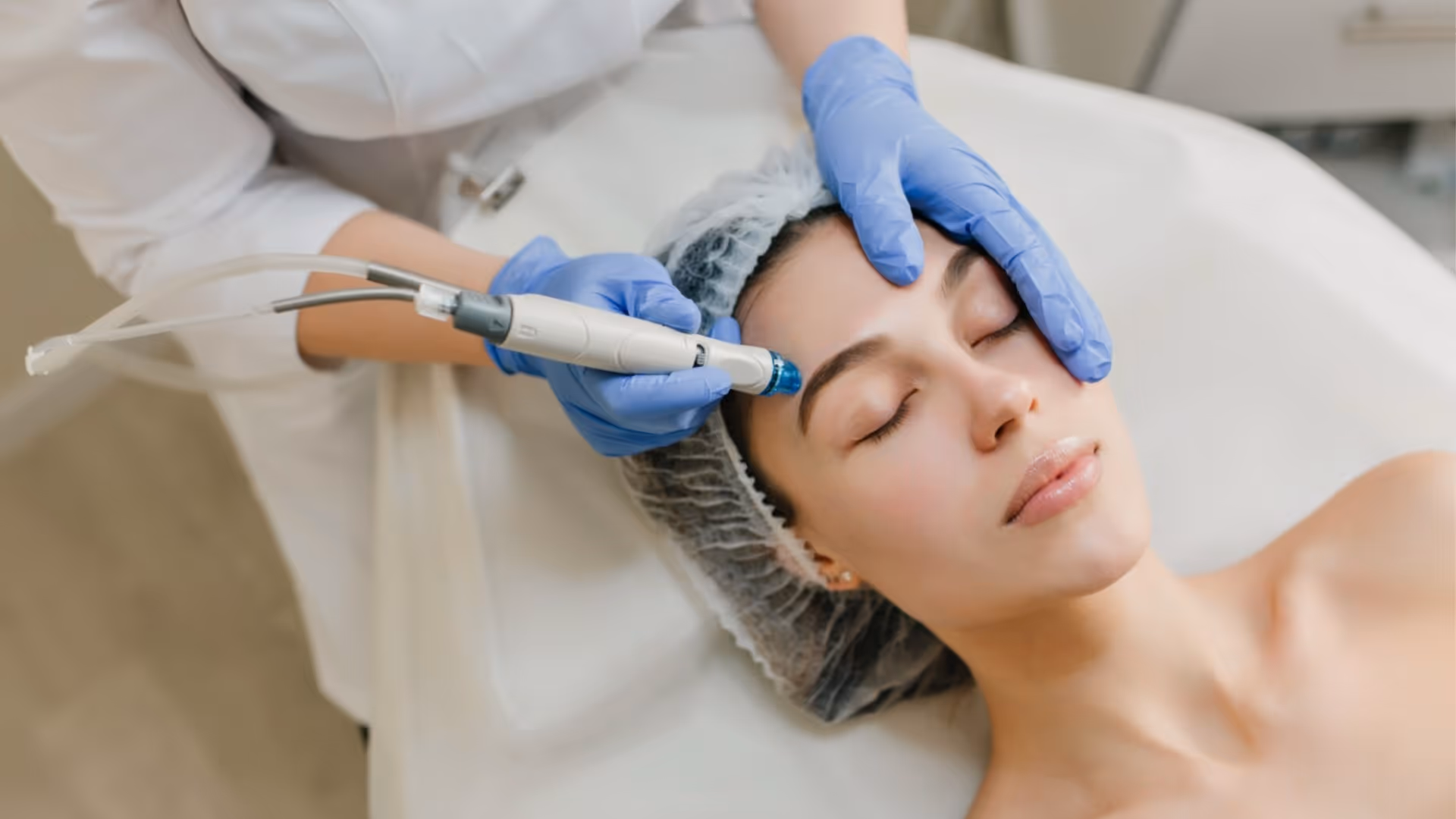 Praticienne avec gants bleus appliquant un dispositif Hydrafacial® sur le front d'une femme allongée avec charlotte, en cabinet esthétique