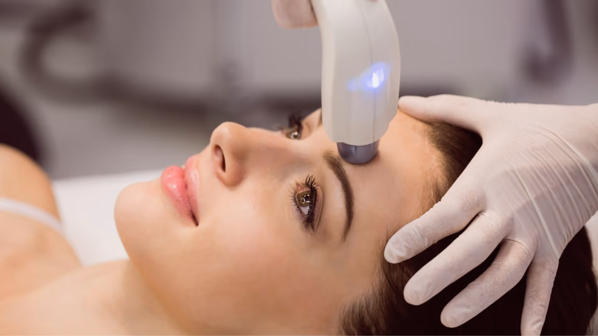 Femme allongée recevant un traitement de photorajeunissement, dispositif blanc émettant une lumière bleue appliqué sur le front, tenu par une main gantée blanche