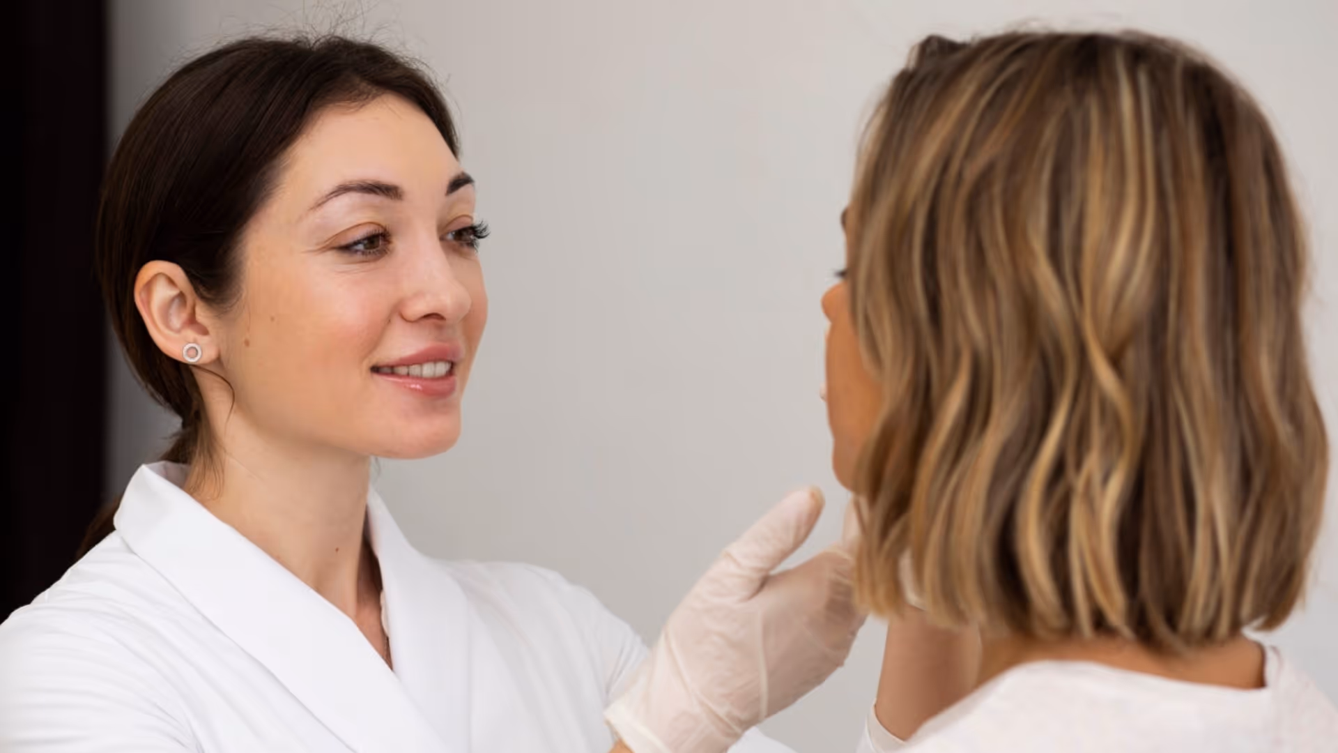 Esthéticienne en blouse blanche examinant le visage d'une cliente aux cheveux blonds courts tout en portant des gants.