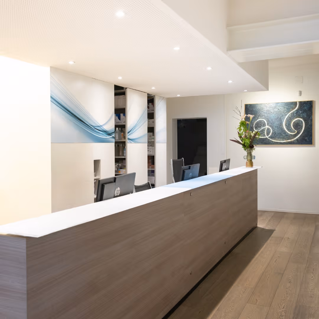 Accueil moderne d'un cabinet avec un comptoir en bois clair, deux ordinateurs, une décoration florale et une œuvre d'art abstraite au mur.