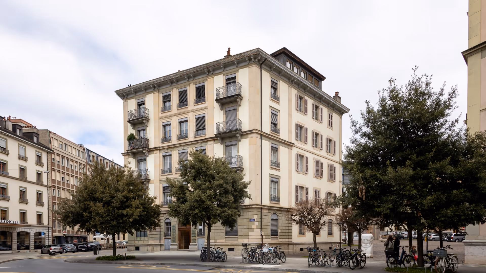 Bâtiment résidentiel de quatre étages avec balcons et volets entouré d'arbres et de vélos stationnés sur un coin de rue urbain.