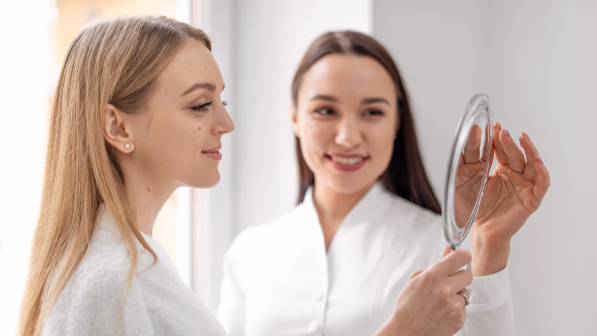 Deux femmes souriantes, l'une tenant une petite glace ronde pendant que l'autre regarde son reflet, en consultation dans un cabinet esthétique.