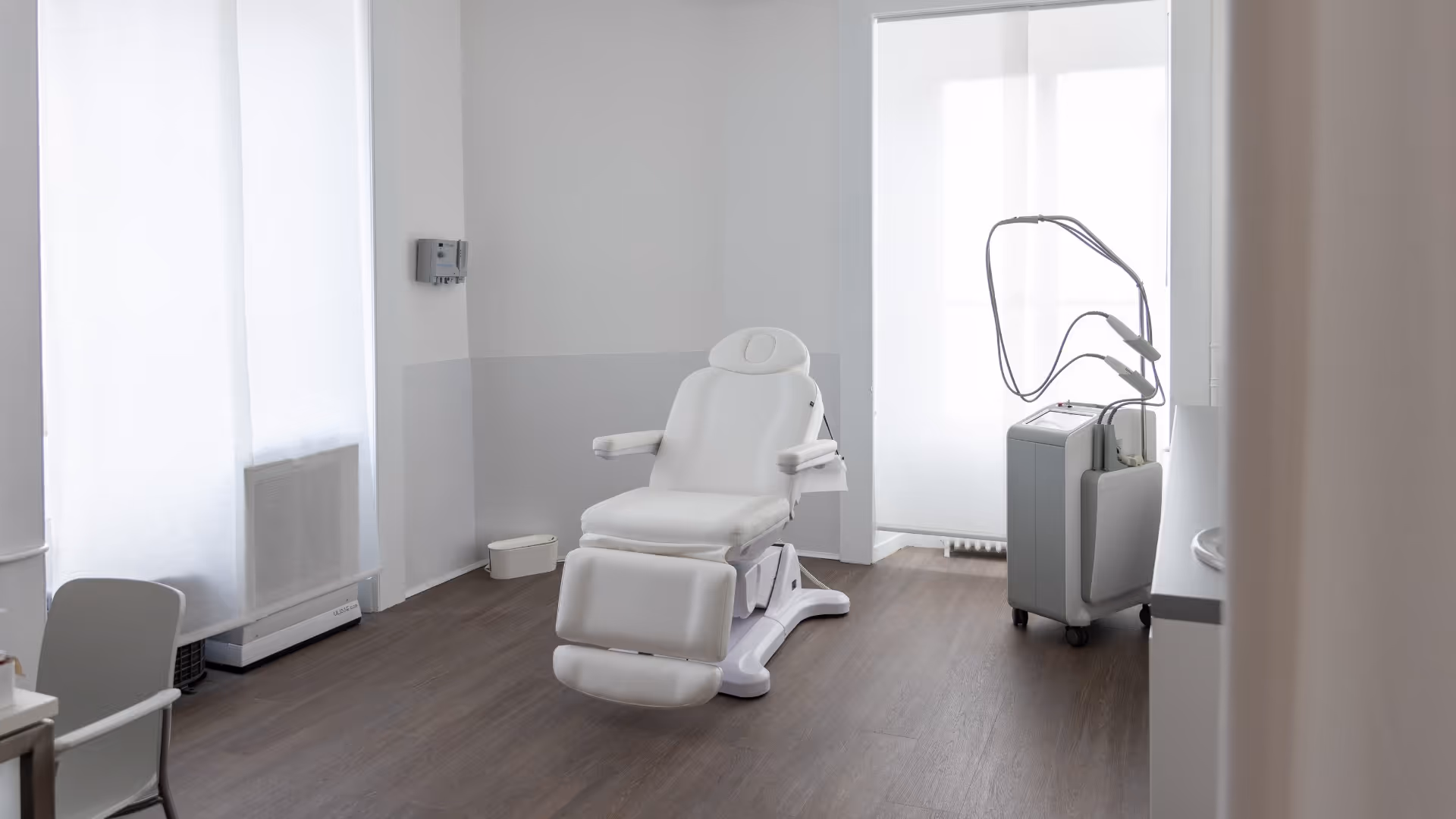 Salle de consultation dermatologique claire avec une chaise médicale blanche ajustable au centre et un appareil médical sur roulettes à côté.
