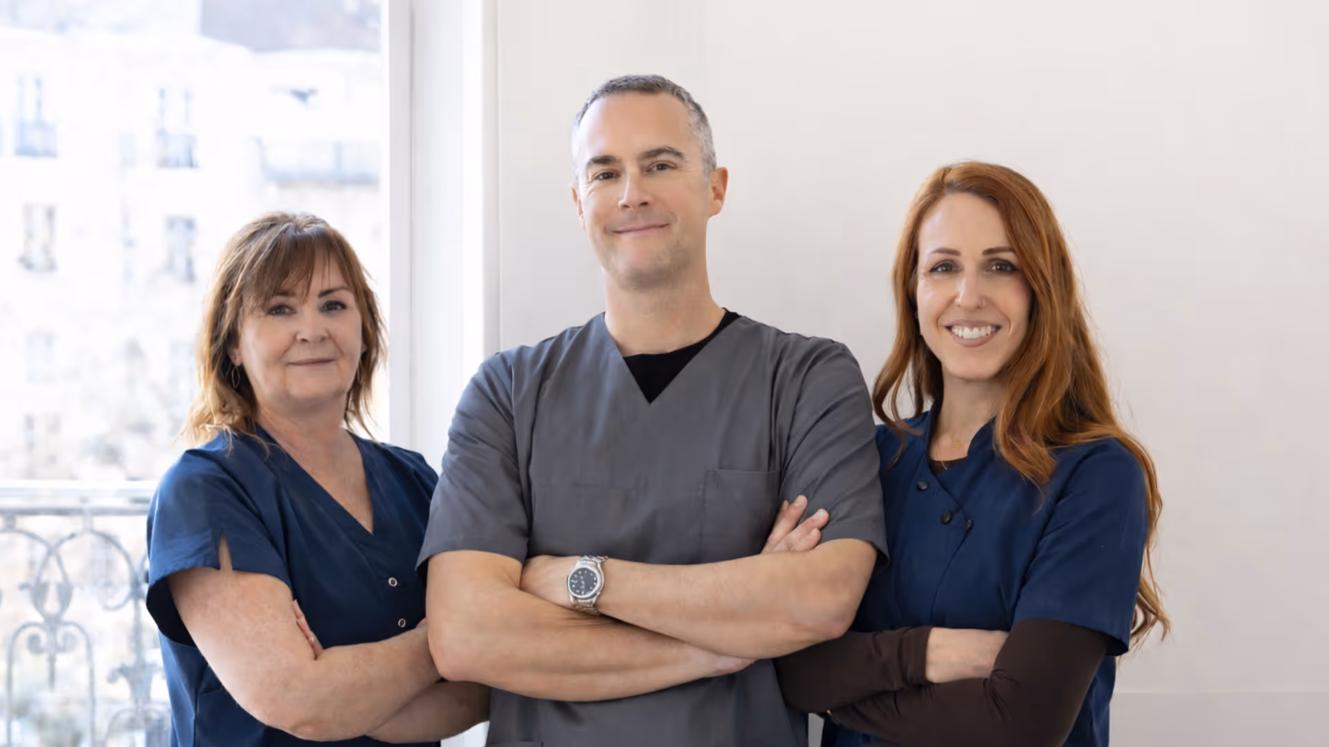 Équipe médicale souriante composée de deux femmes et un homme portant des blouses médicales, bras croisés, devant une fenêtre.