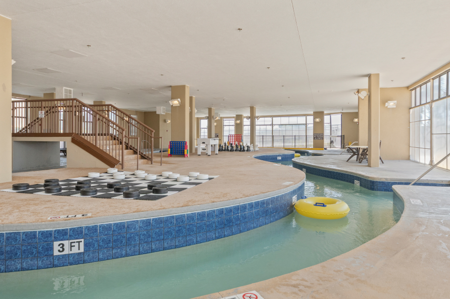 Indoor resort pool area with a lazy river, hot tub, giant checkers board, Connect Four game, and large windows at a North Myrtle Beach vacation rental.