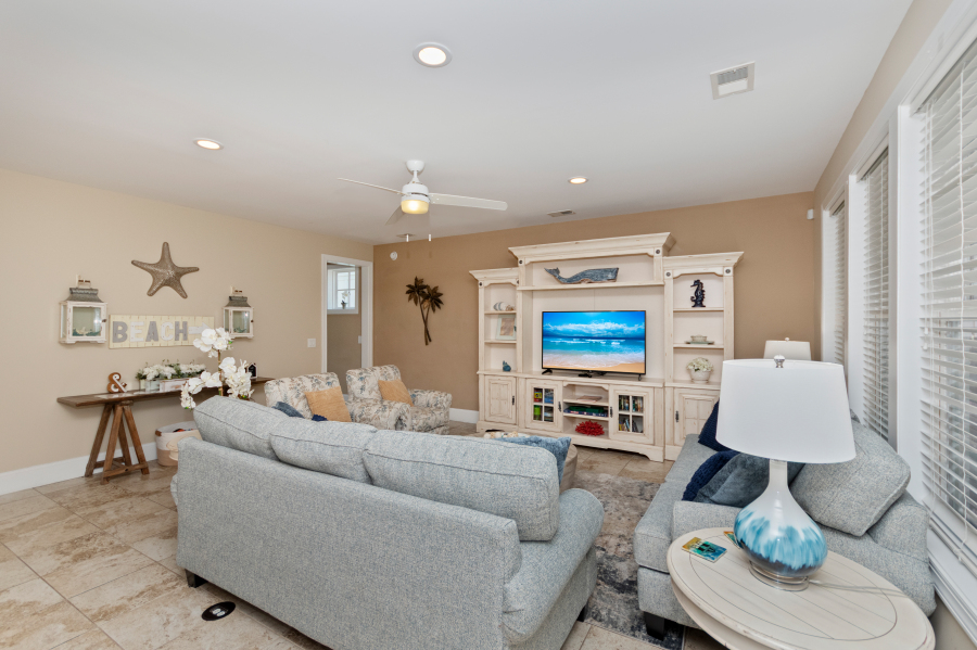 Coastal living room with gray sofas, beach-themed decor, TV entertainment center, ceiling fan, and large windows at a North Myrtle Beach vacation rental.