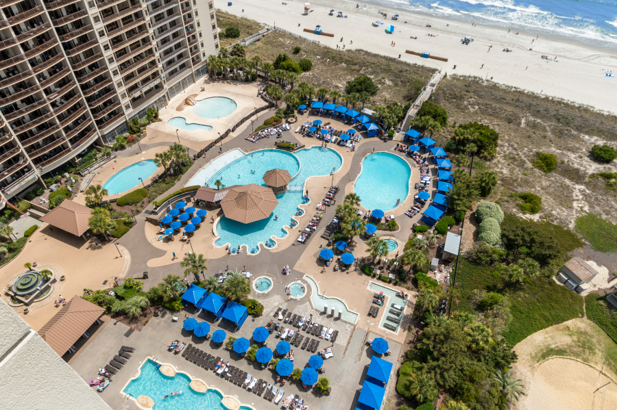 Aerial view of oceanfront resort pools, blue umbrellas, lounge chairs, and beach access along the North Myrtle Beach shoreline.
