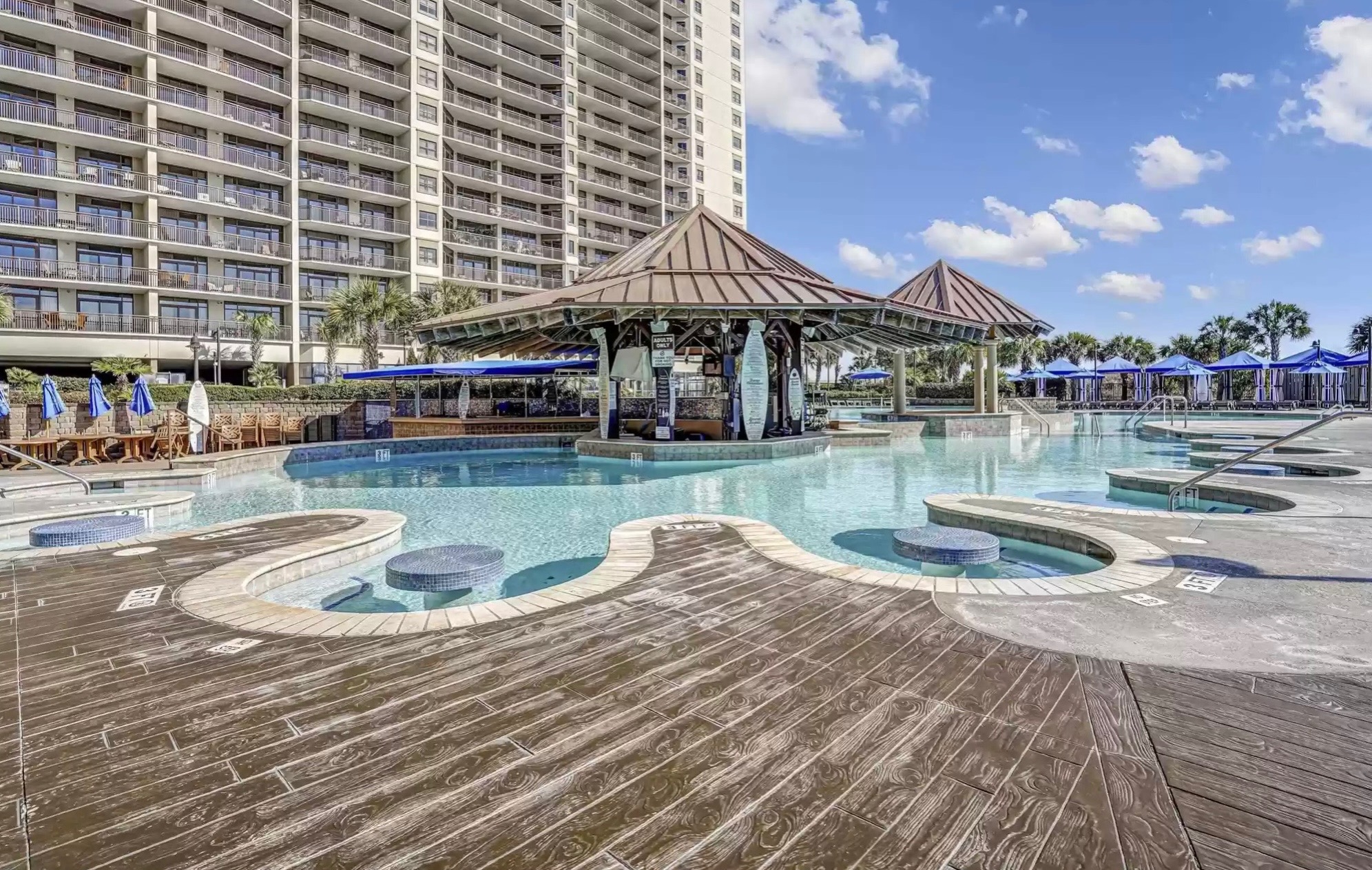 Resort pool area with a swim-up bar, shallow lounge seating, hot tubs, blue cabanas, and a high-rise oceanfront building in the background.