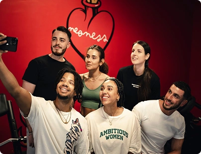 Groupe de cinq jeunes adultes souriants prenant un selfie devant un mur rouge avec un dessin de cœur et le mot « meoness ».