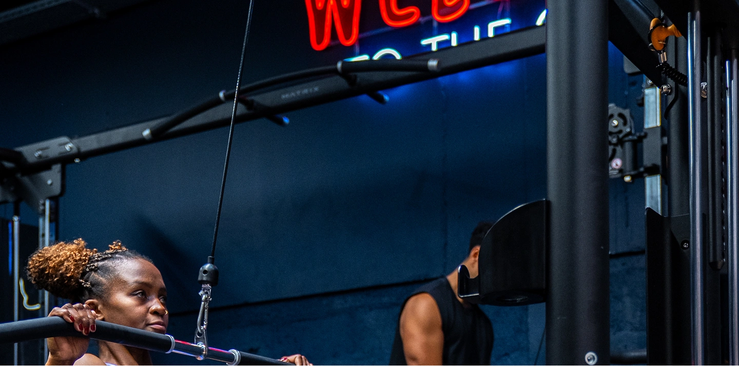 Femme faisant un exercice de tirage à la poulie haute dans une salle de sport avec un homme en arrière-plan.