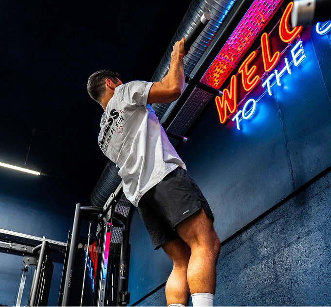 Homme effectuant une traction dans une salle de sport avec un mur bleu et une enseigne au néon colorée.
