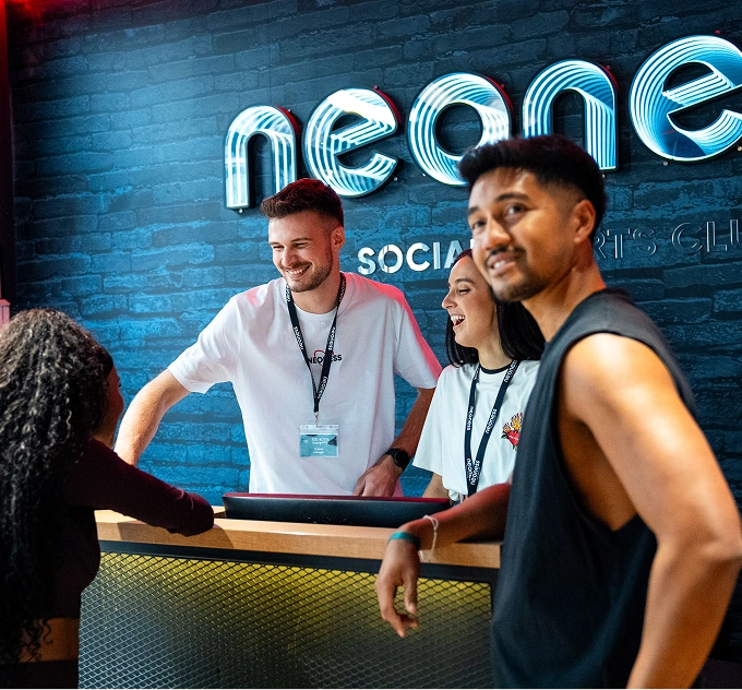 Quatre jeunes adultes souriants discutent dans un club social sportif avec un mur en briques et enseigne lumineuse néon en arrière-plan.