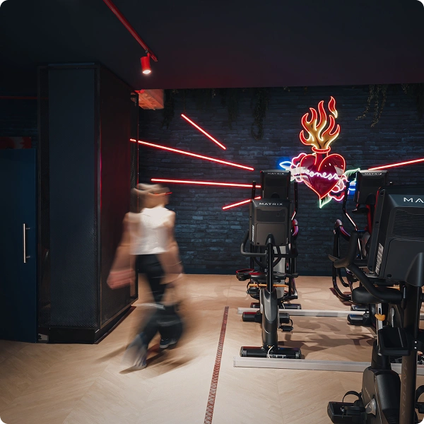 Personne en mouvement dans une salle de sport avec des vélos d'appartement alignés et un mur noir décoré d'un néon en forme de cœur enflammé.