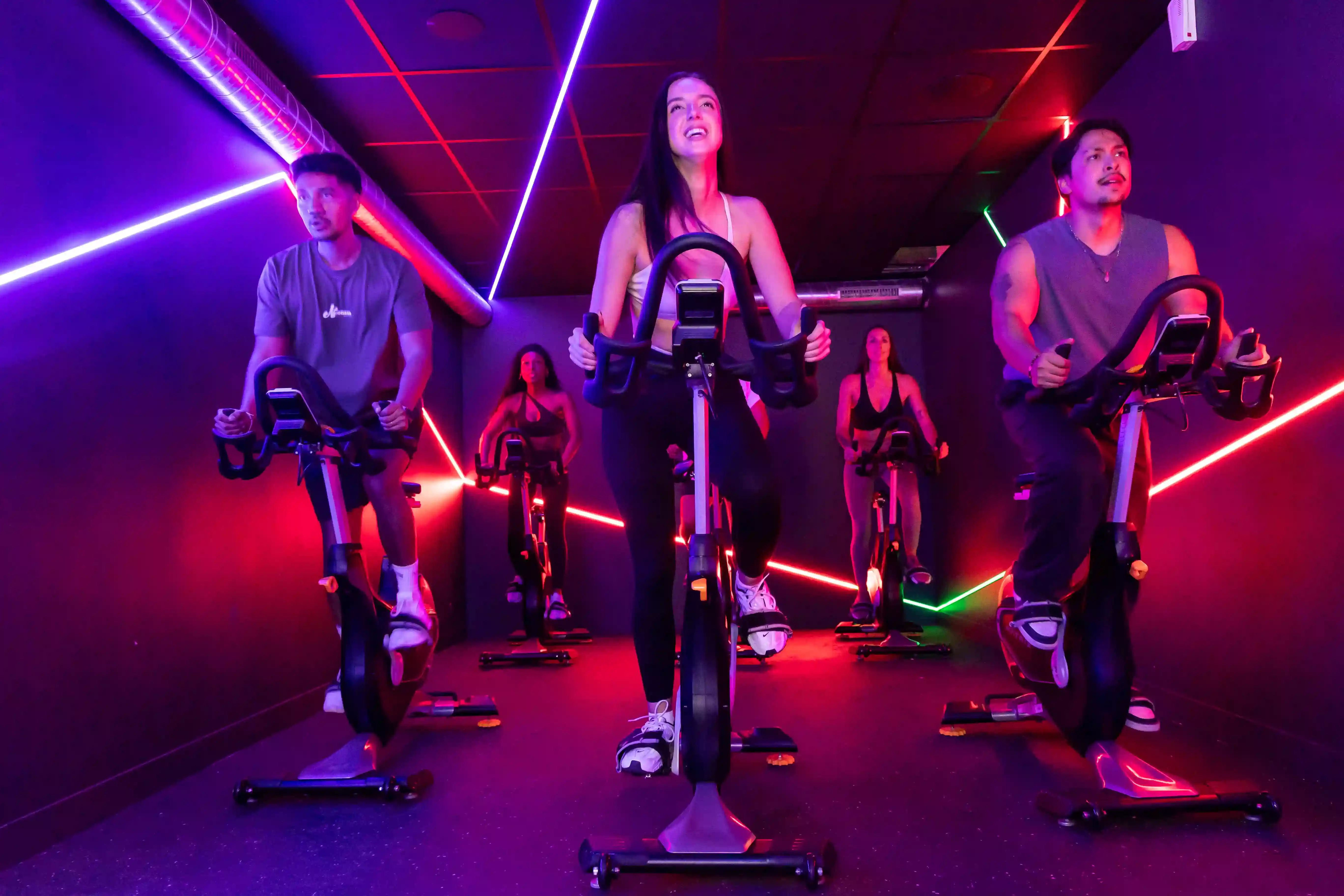 Groupe de personnes faisant du vélo en salle dans une pièce sombre éclairée par des lumières LED colorées.