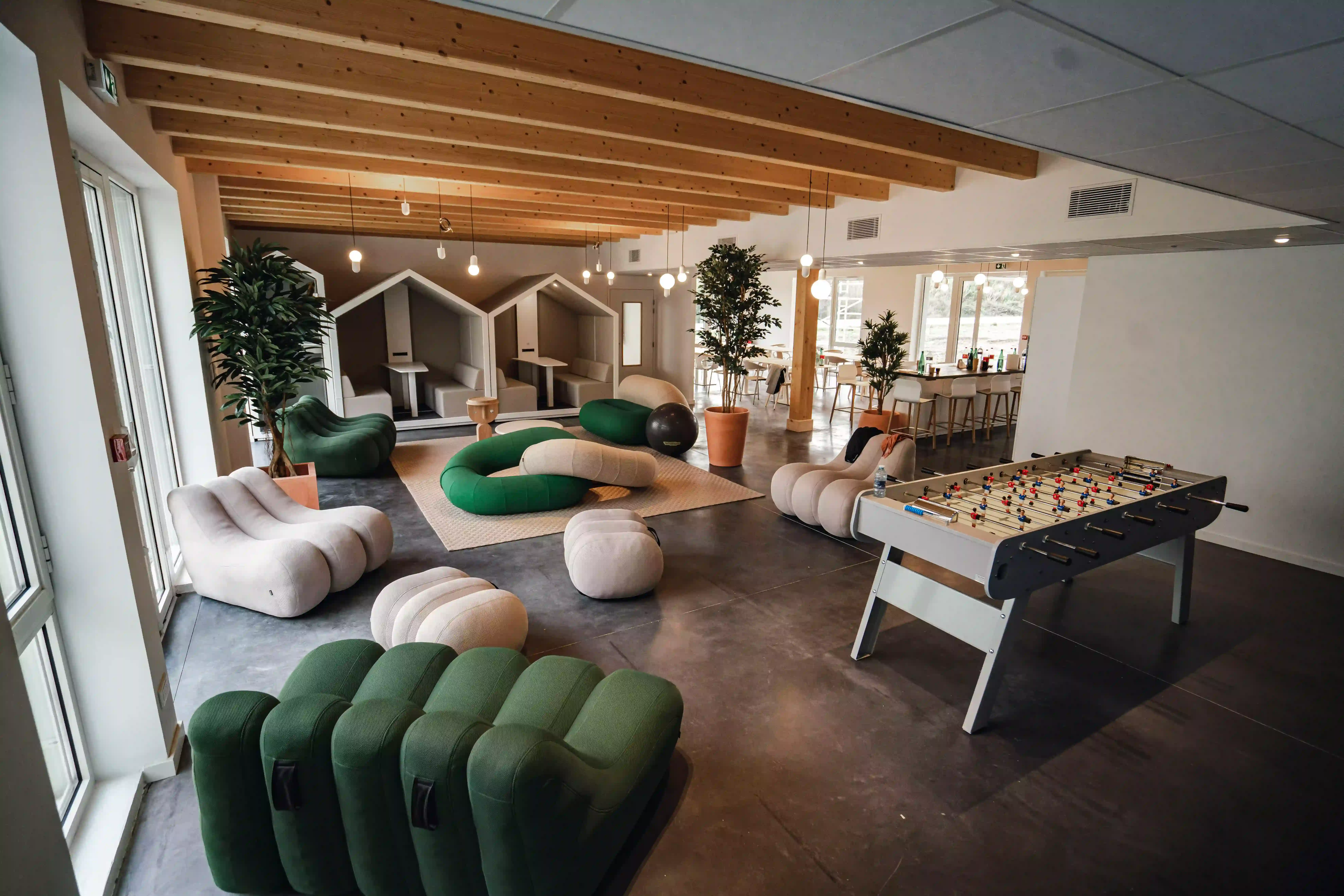 Salle de détente moderne avec meubles modulaires verts et beiges, baby-foot, et espaces de réunion en alcôve sous poutres en bois apparentes.