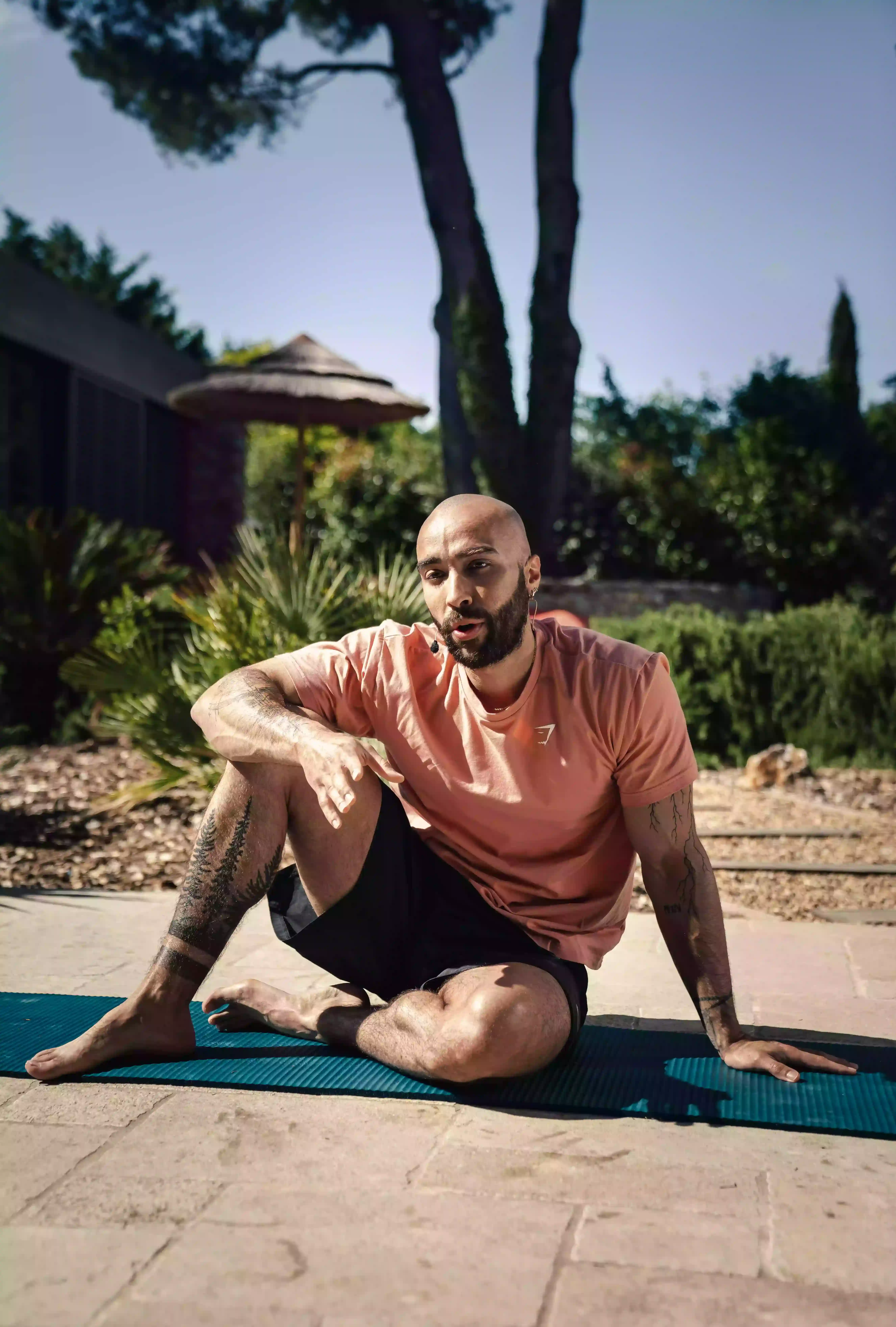 Homme assis sur un tapis de yoga en plein air, portant un t-shirt saumon et un short noir, entouré de végétation.