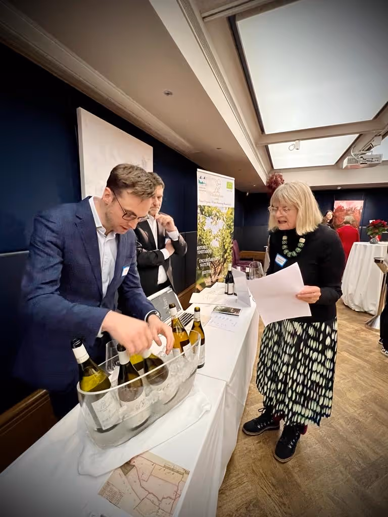 Man in suit handling wine bottles at a table while a woman with papers stands nearby viewing him.