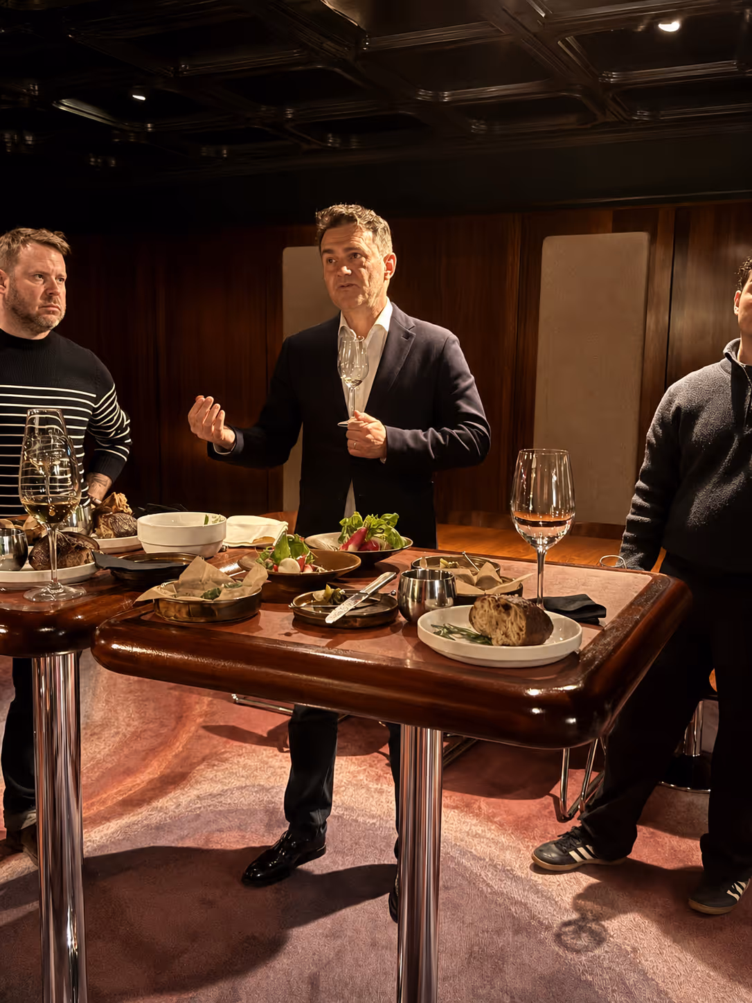 Three men standing around a table with food and wine glasses in a dimly lit room.