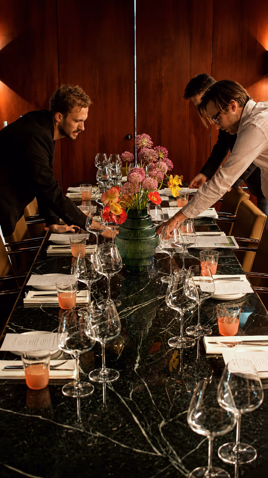 Three men setting a formal dining table with wine glasses and a central green vase of flowers.