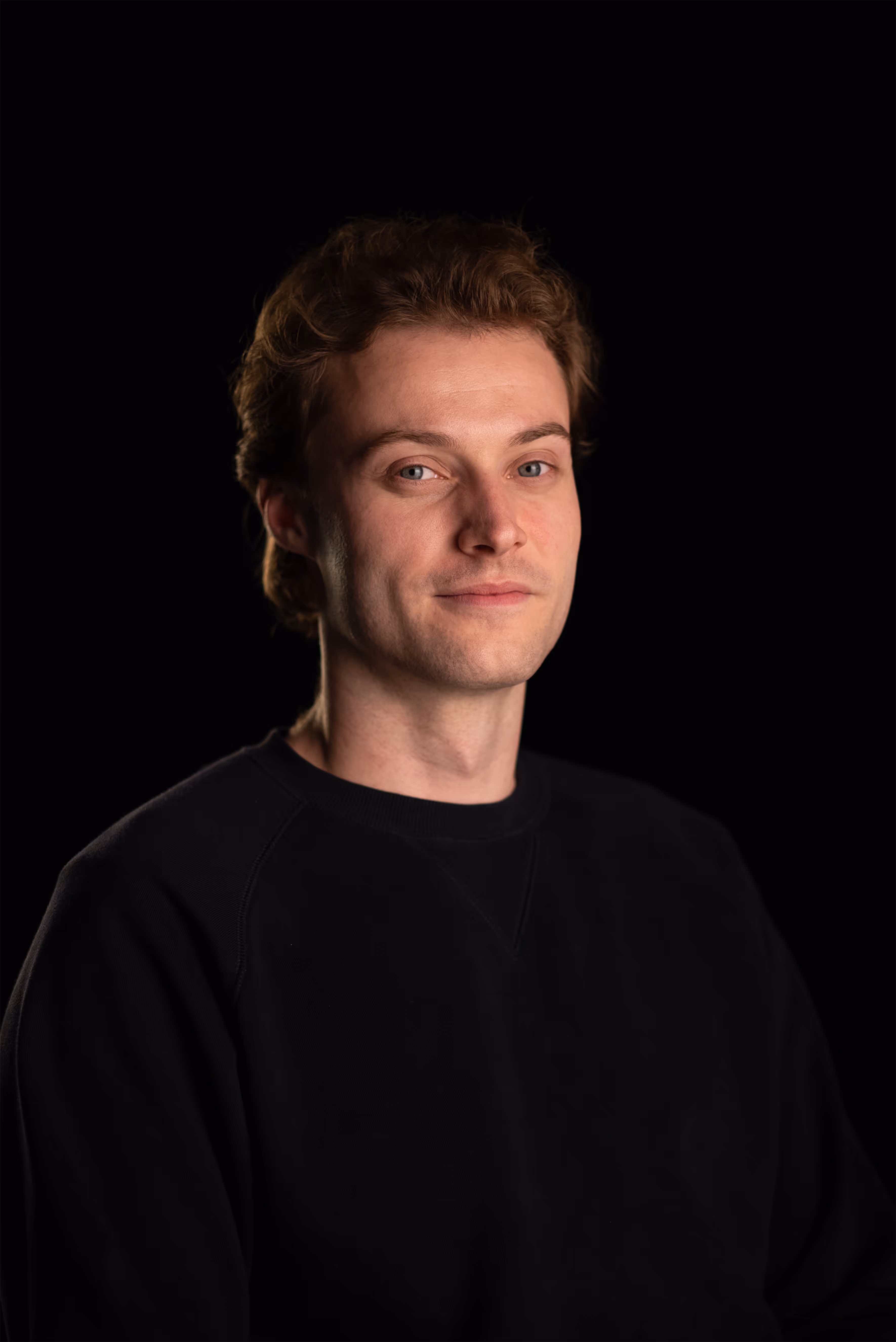 Jake Greig. Young man with light skin and curly brown hair wearing a black shirt against black background.