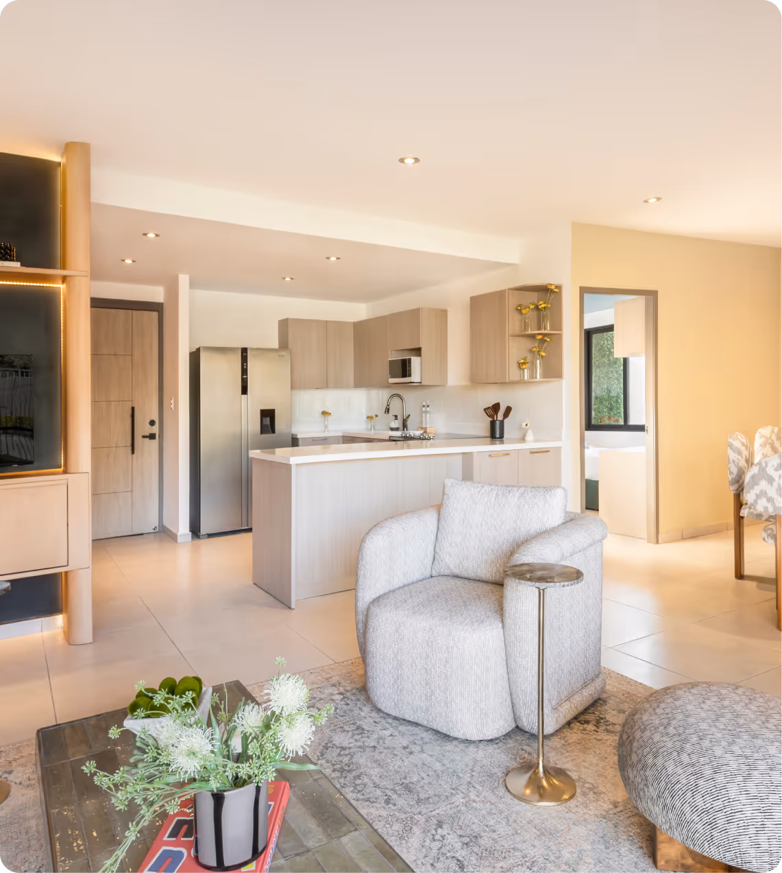 Modern open living room and kitchen with a gray armchair, small side table, flower vase on coffee table, and light wood cabinetry.