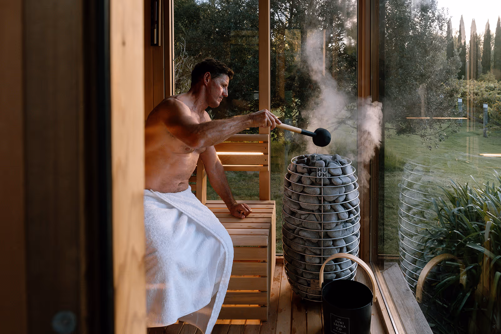 Man sitting in a sauna pouring water onto the heater, creating steam
