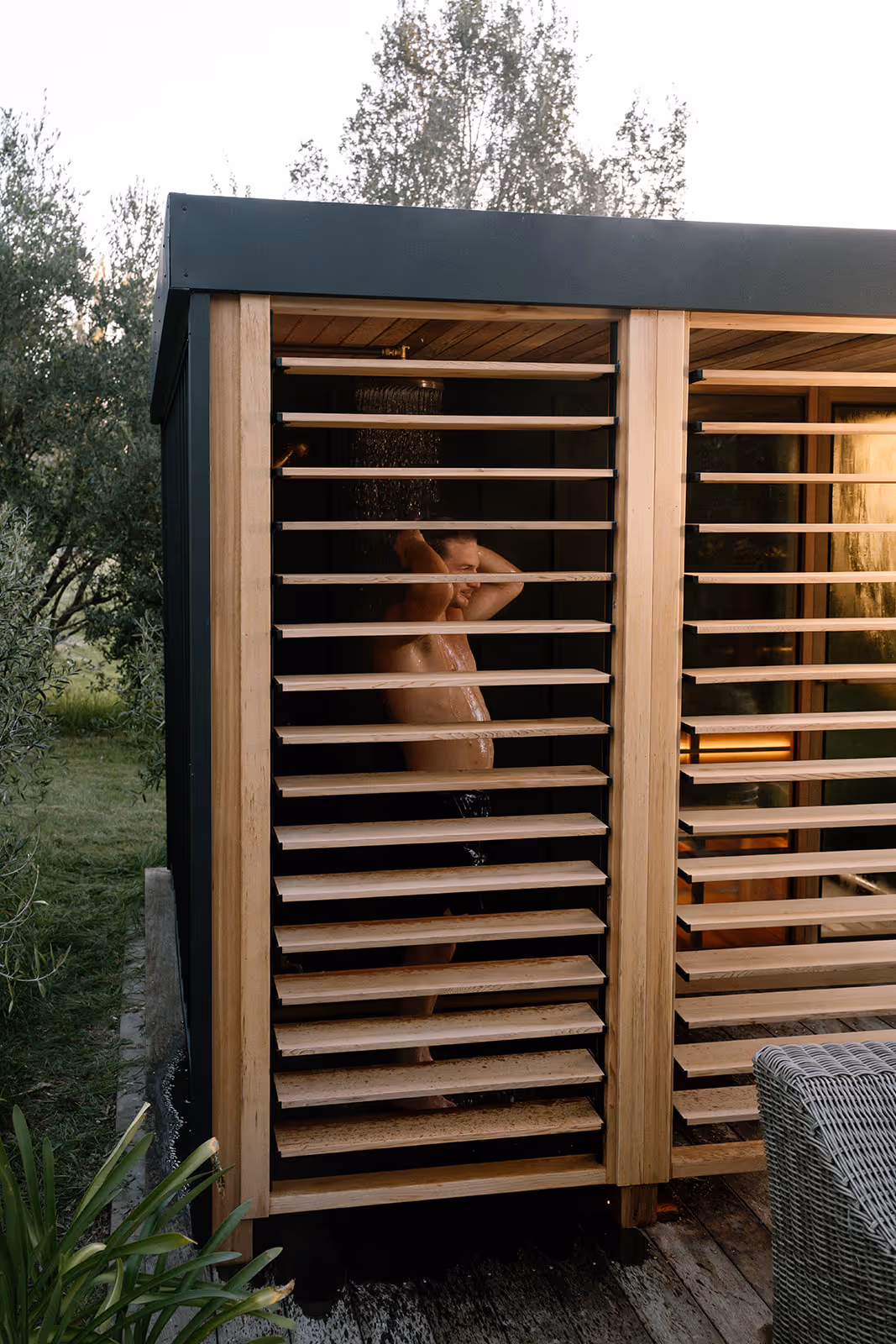 Man taking an outdoor shower inside a wooden slatted enclosure surrounded by greenery.