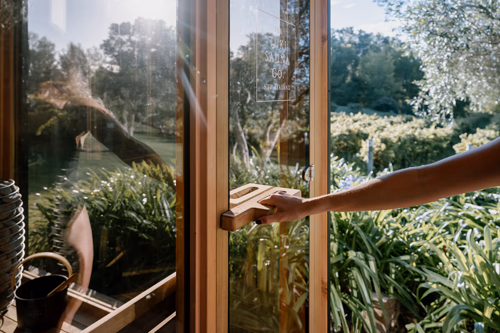 Girl holding the handle of a glass sauna door with floor-to-ceiling panels, branded The Tuki Sauna Co