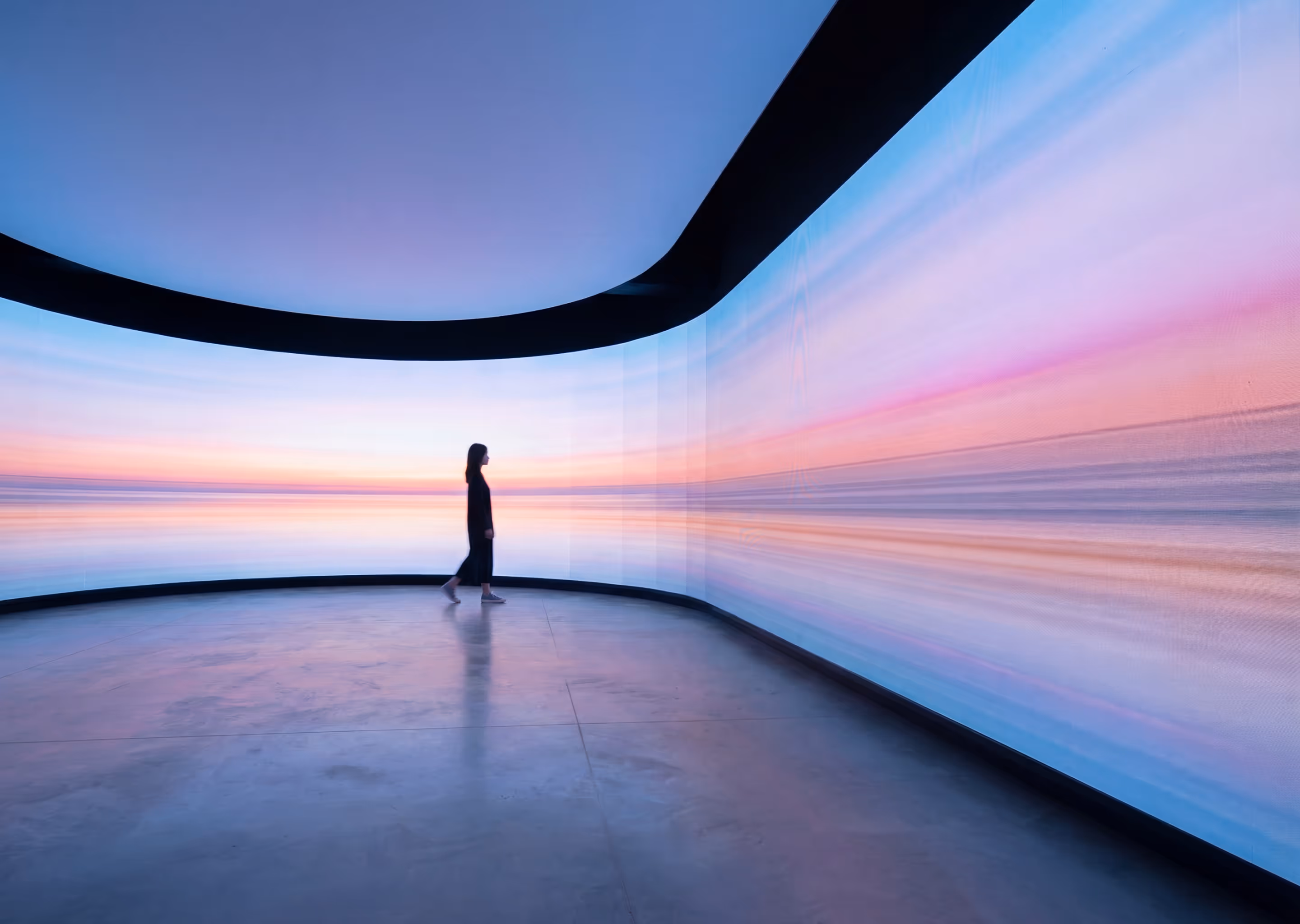 Person standing in a modern room with curved walls displaying a digital sunset landscape with pink, blue, and purple hues.