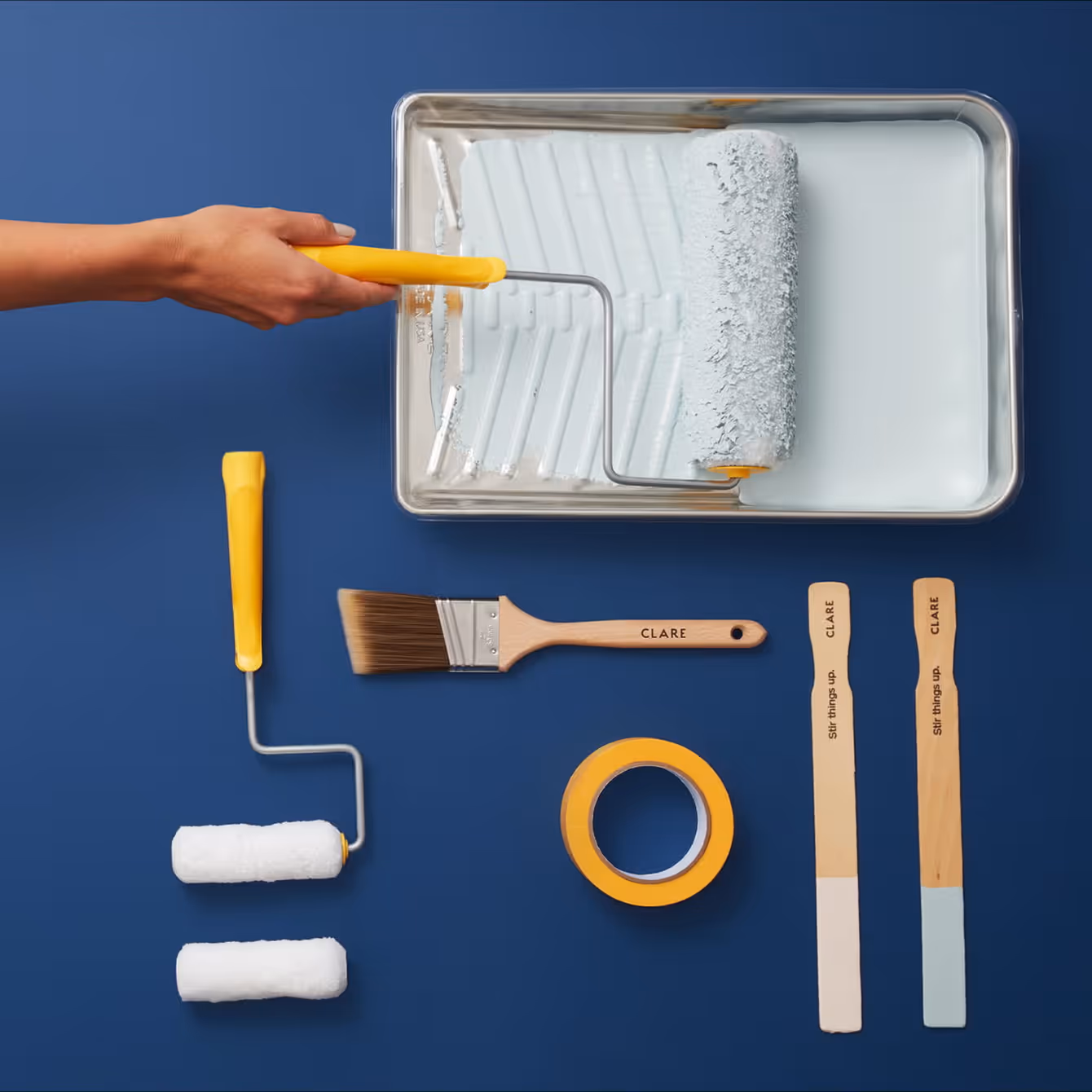 Painting tools including a paint roller dipped in white paint, two additional rollers, a paintbrush labeled Clare, a roll of yellow painter's tape, and two wooden stir sticks with paint samples on a blue background.