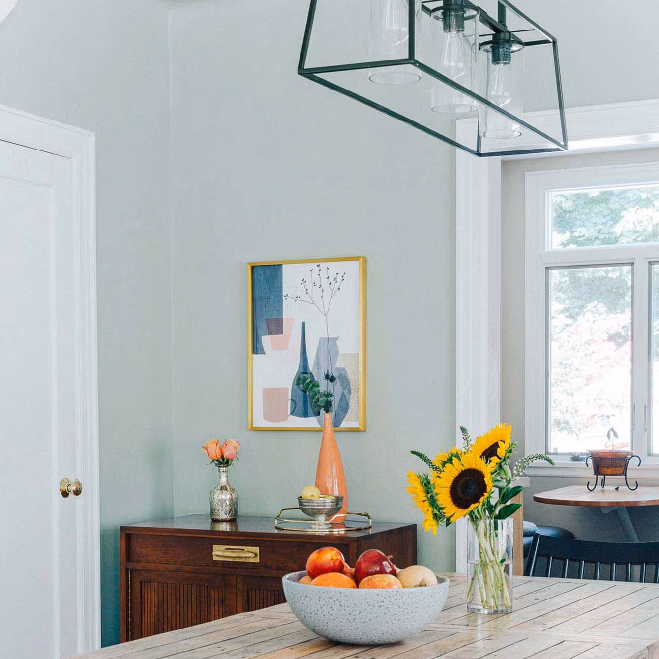 Sunflowers in a glass vase and a bowl of fruit on a wooden table in a bright dining room with a sideboard, decorative vases, and abstract wall art.