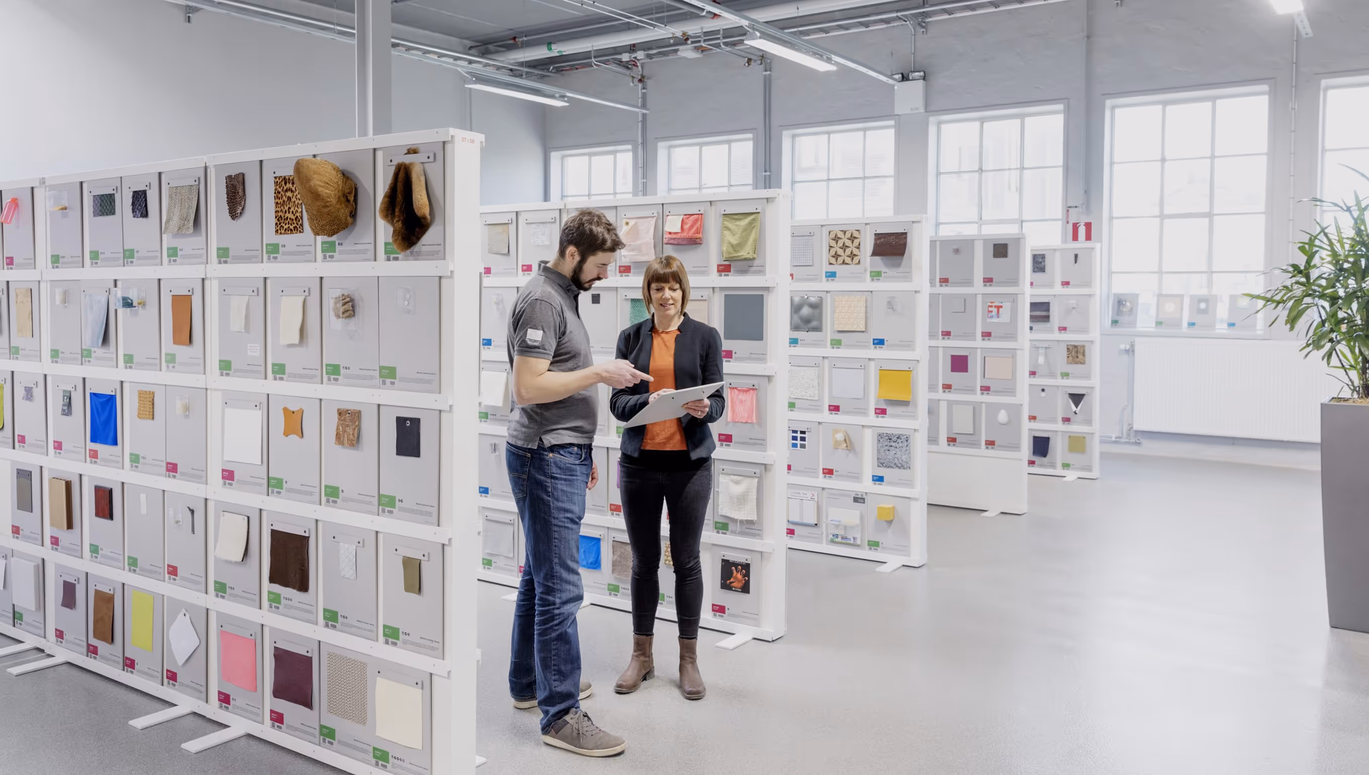 Two people discussing with a clipboard in a bright room filled with panels displaying various fabric and material samples.