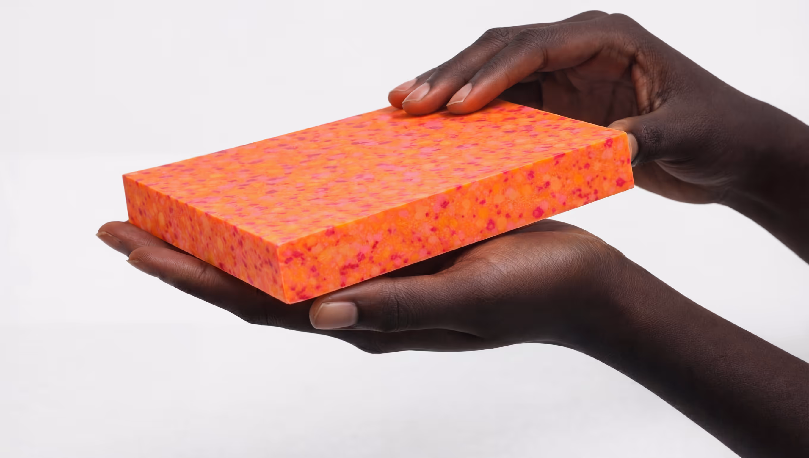 Dark-skinned hands holding a rectangular orange and pink speckled foam block.