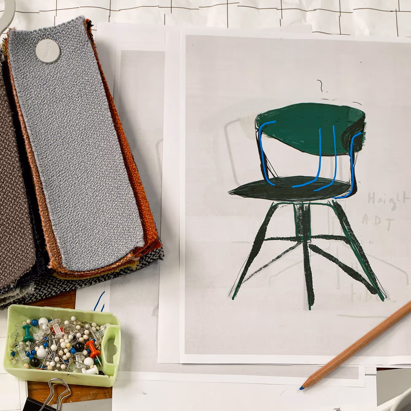 Desk with fabric swatches, sewing pins, a pencil, and a printed sketch of a modern green chair design.