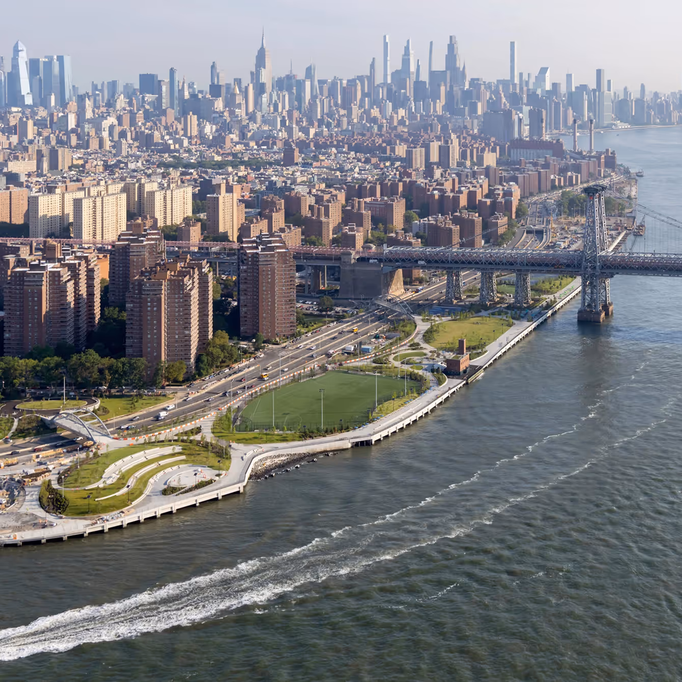 Aerial view of the Williamsburg Bridge crossing the East River with Manhattan skyline in the background and a waterfront park in the foreground.
