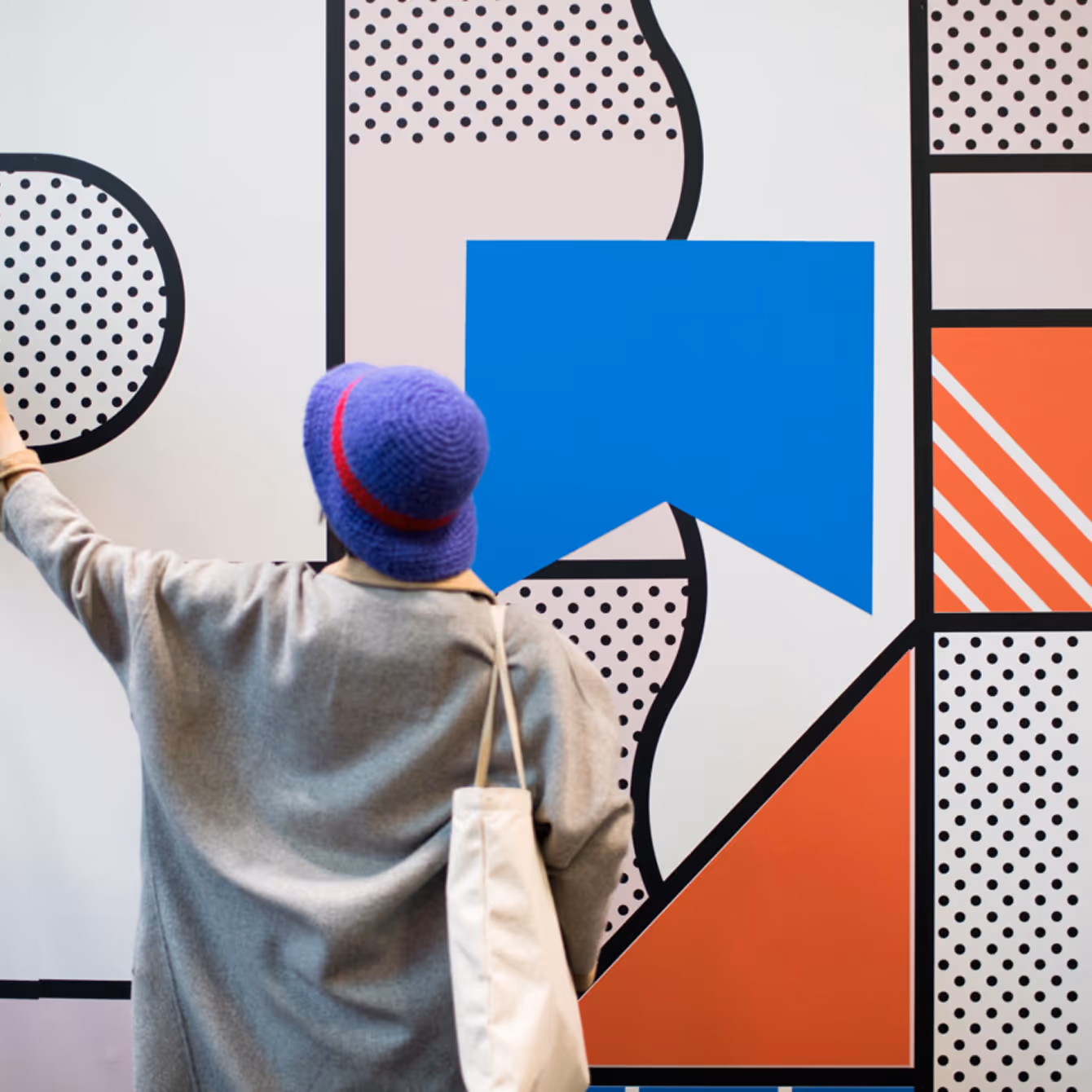 Person wearing a blue hat and gray coat reaching towards a colorful wall mural with geometric shapes and patterns.