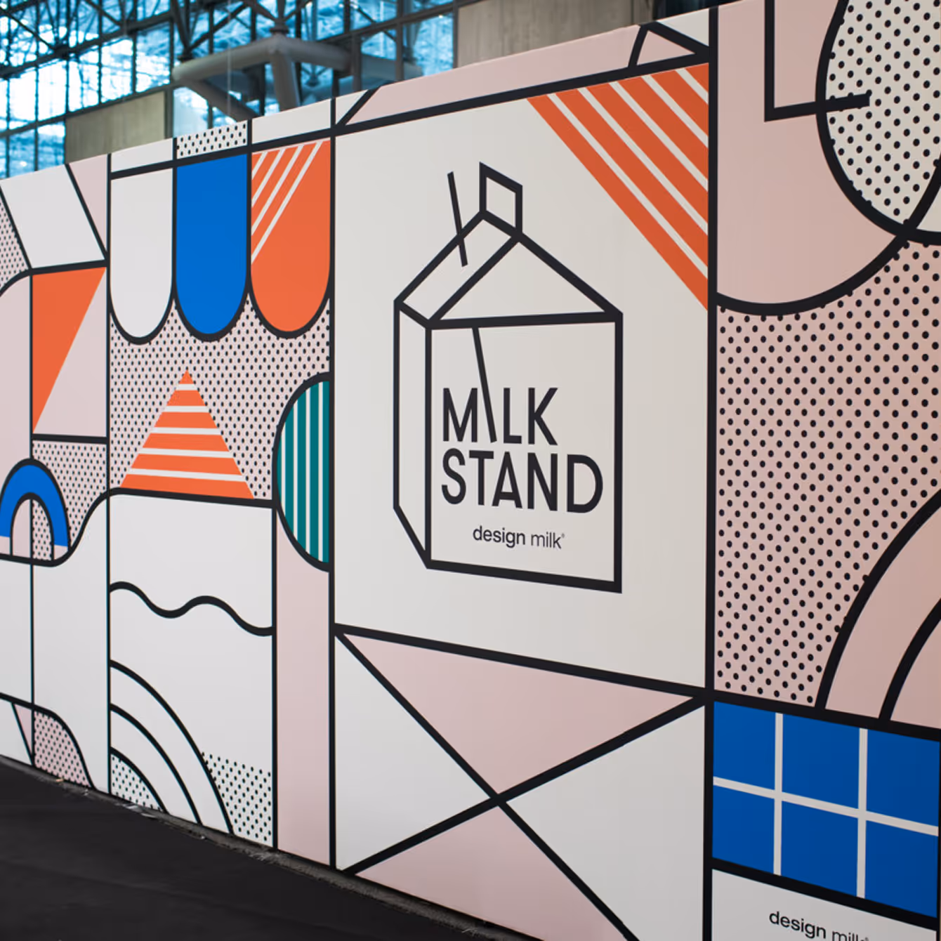 Colorful geometric mural with a graphic of a milk carton labeled 'MLK STAND design milk' inside a building with glass ceiling.