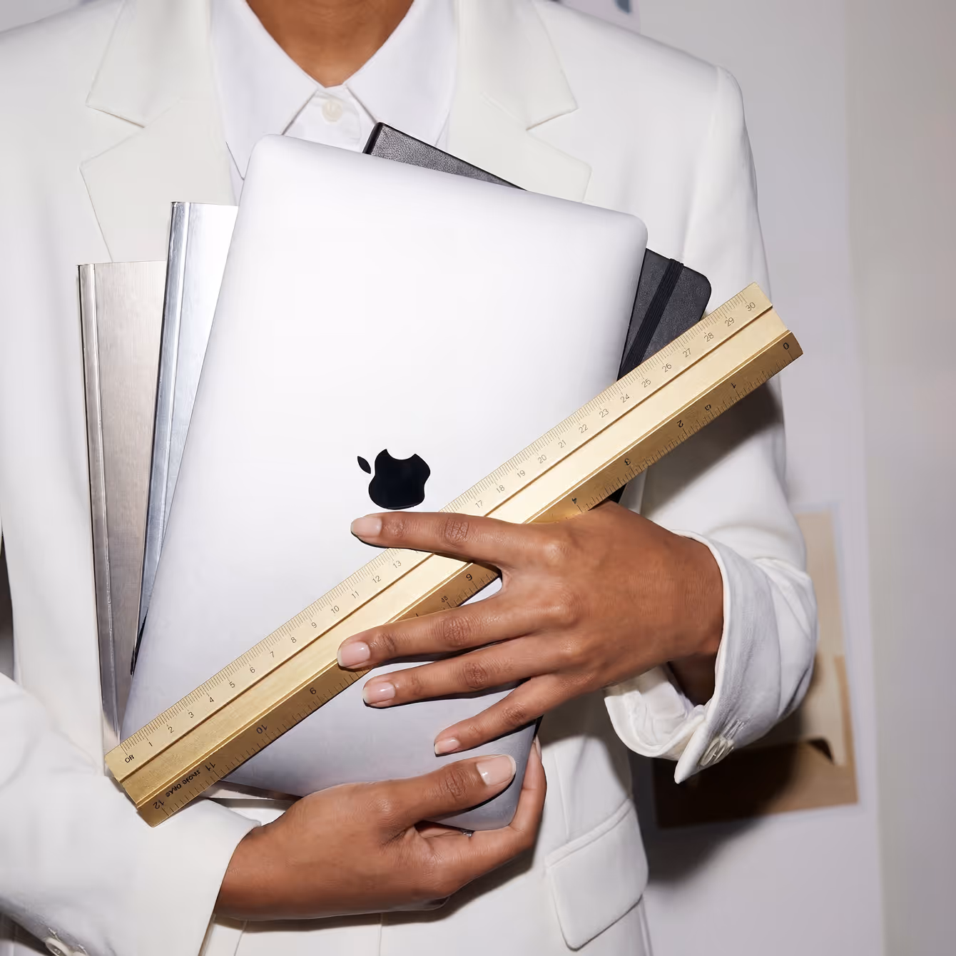 Person in white blazer holding an Apple laptop, notebooks, and a wooden ruler.