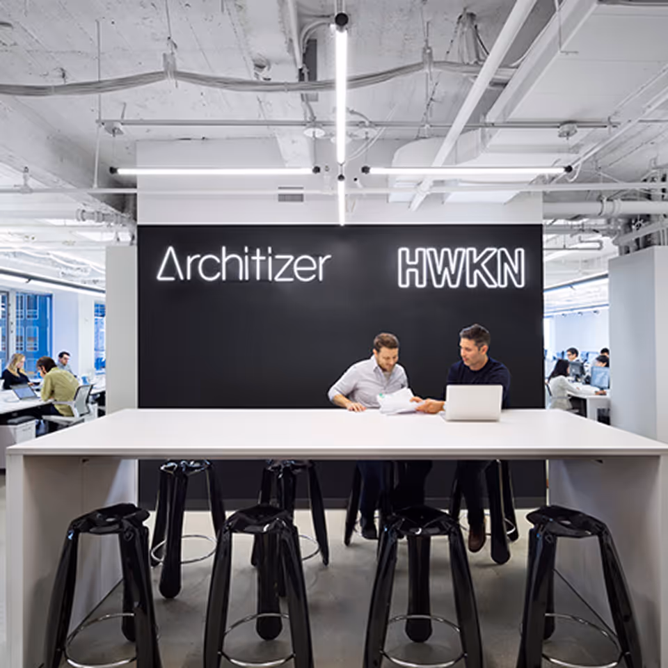 Two men working at a white table with a laptop and papers in a modern office with Architizer and HWKN neon signs on the black wall behind them.